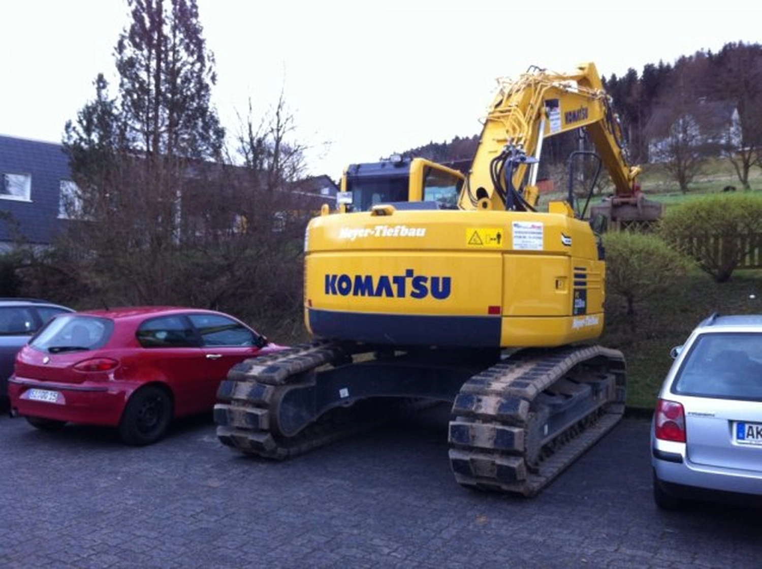 Hans Werner Meyer GmbH - Tiefbauunternehmen - Ein großer gelber Komatsu-Bagger parkte auf einem Parkplatz zwischen einem roten Schrägheckwagen und einem silbernen Kombi.