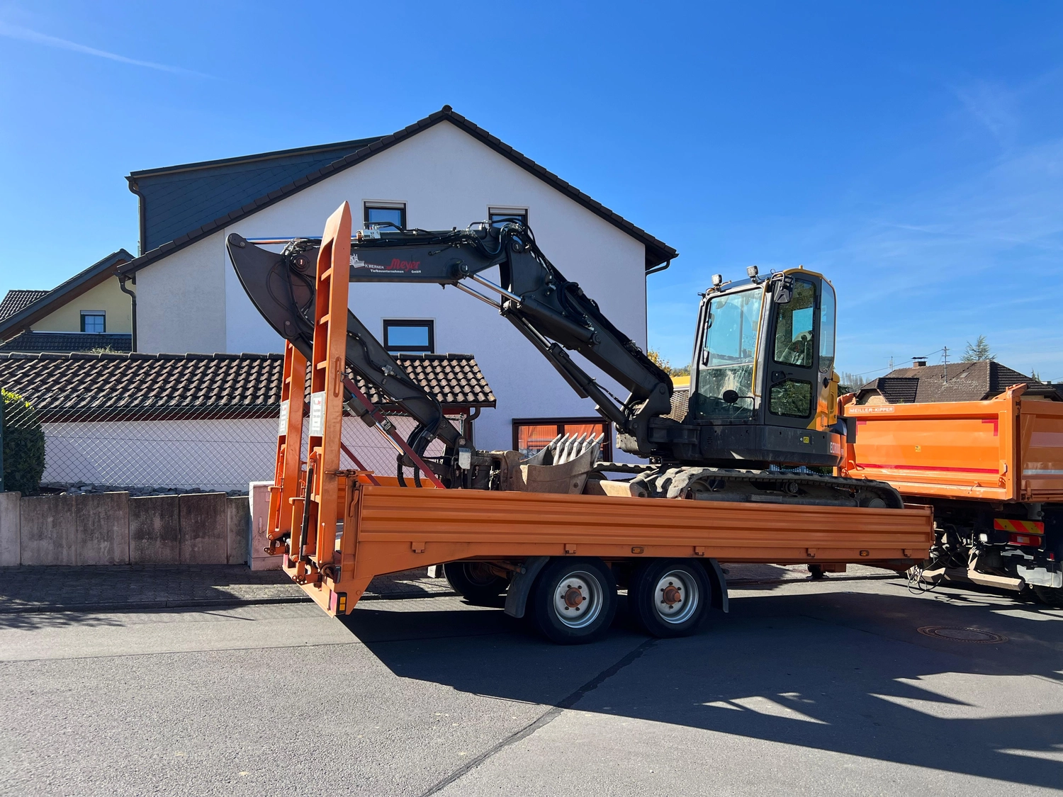 Hans Werner Meyer GmbH - Tiefbauunternehmen - Ein Bagger, der auf einem Anhänger durch eine sonnige Straße transportiert wird.