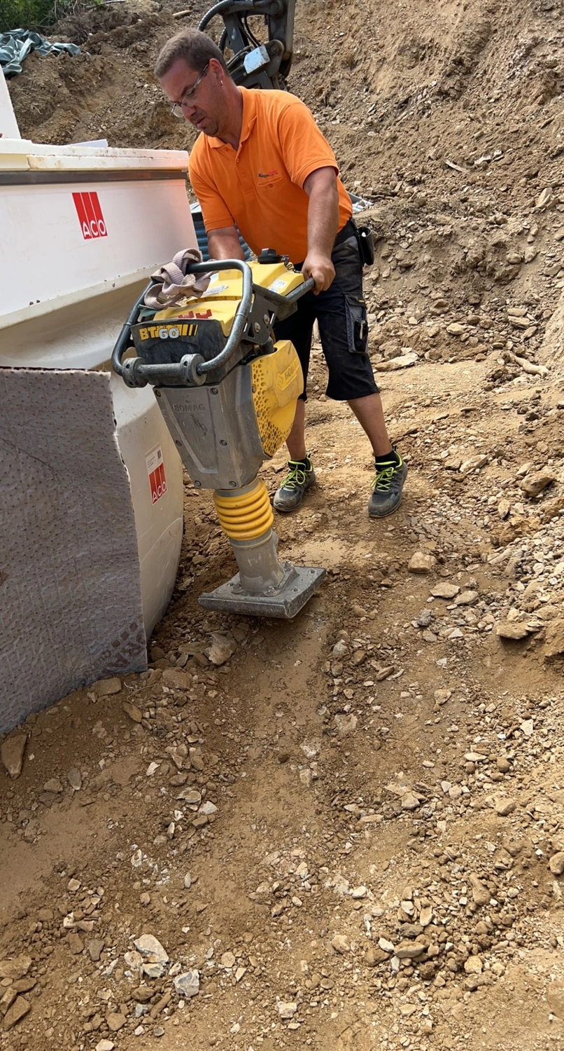 Hans Werner Meyer GmbH - Tiefbauunternehmen - Ein Mann in orangefarbenem Poloshirt und Arbeitskleidung bedient einen gelben Vibrationsstampfer, um den Boden auf einer Baustelle zu verdichten.