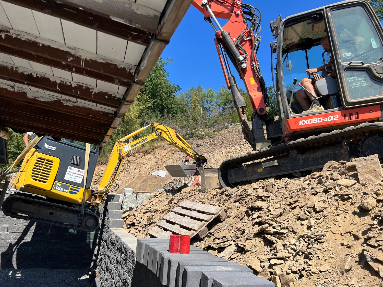 Hans Werner Meyer GmbH - Tiefbauunternehmen - Zwei Bagger, einer gelb und einer orange, arbeiten an einem felsigen Hang neben einer Steinmauer.