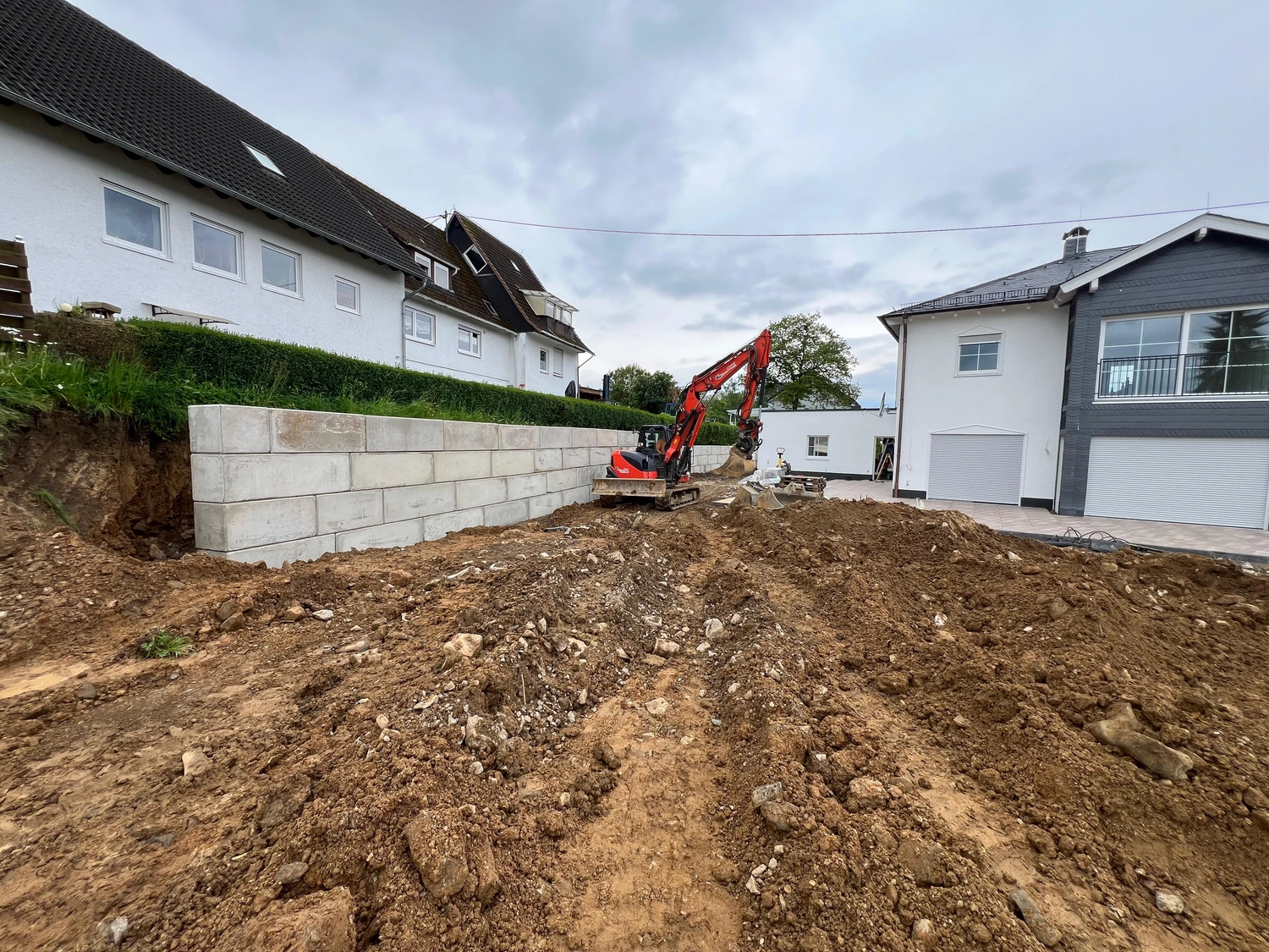 Hans Werner Meyer GmbH - Tiefbauunternehmen - Auf einer Baustelle zwischen zwei Wohnhäusern ist ein roter Bagger im Einsatz. Dort wurde an einem grasbewachsenen Hang eine neue Stützmauer aus Betonblöcken errichtet.