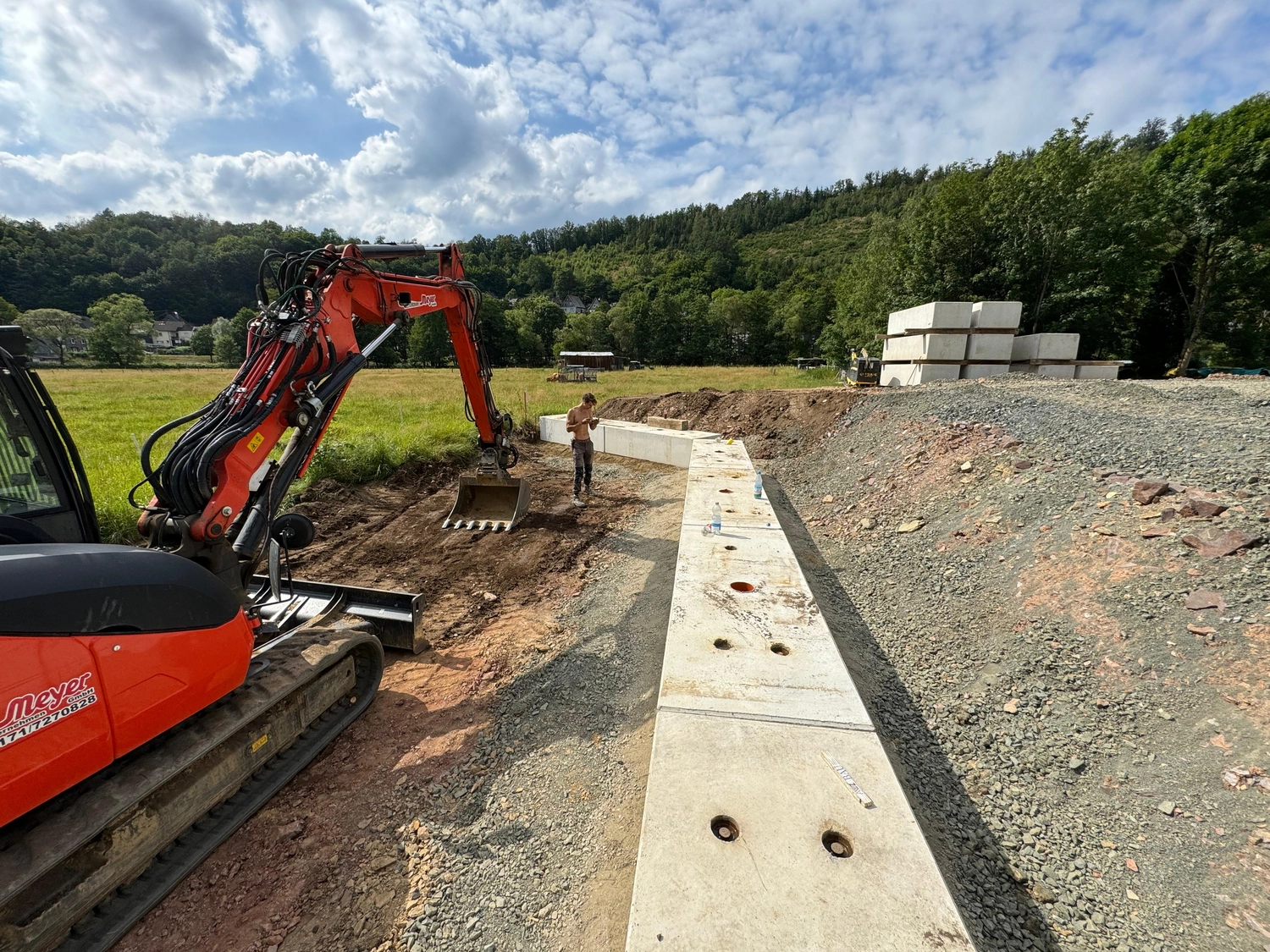 Hans Werner Meyer GmbH - Tiefbauunternehmen - Ein orangefarbener Bagger auf einer Baustelle neben einer neu errichteten Stützmauer aus Betonsteinen.