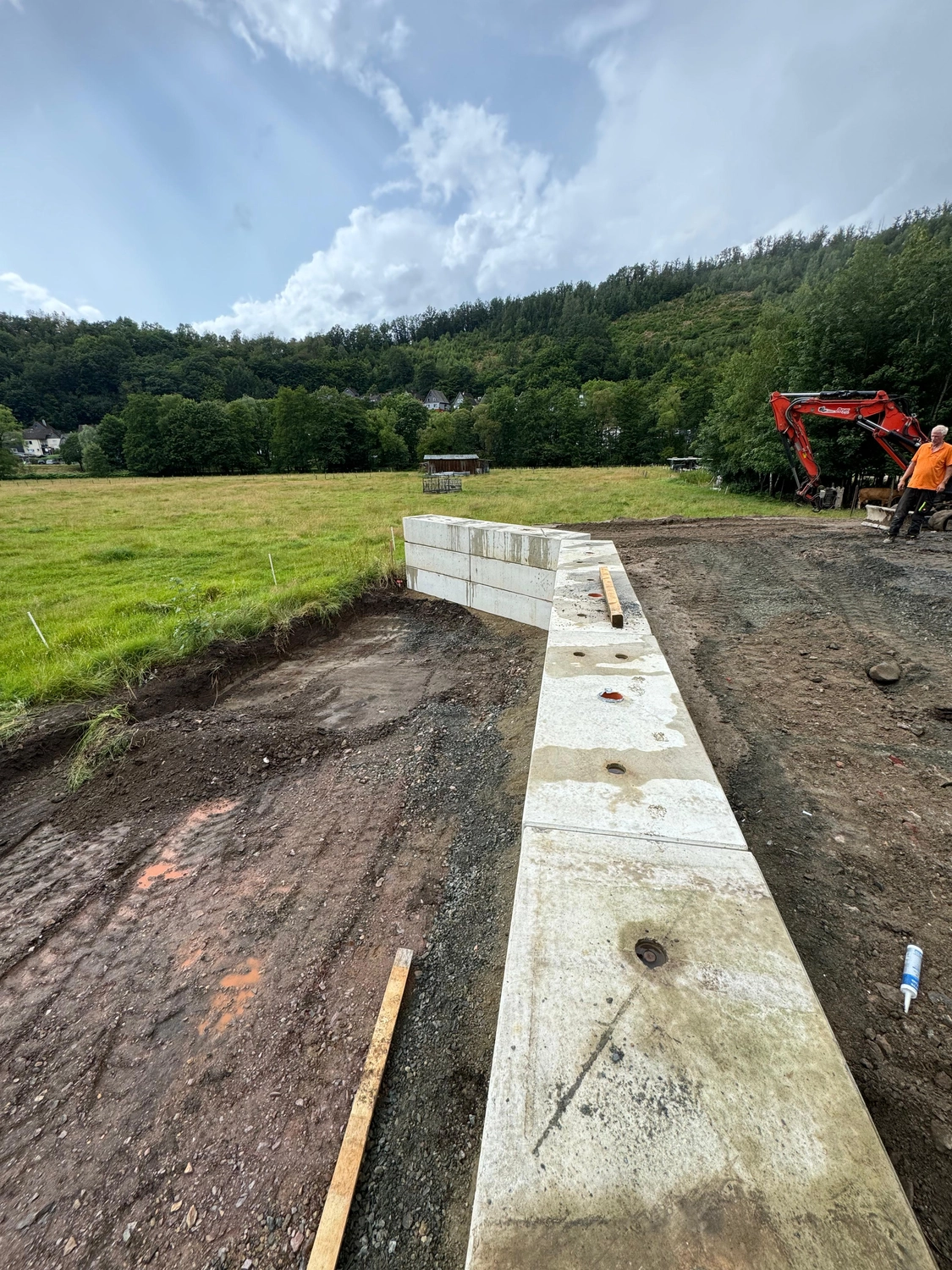 Hans Werner Meyer GmbH - Tiefbauunternehmen - Auf einer Baustelle ist eine teilweise errichtete Stützmauer aus großen, hellen Betonblöcken zu sehen, die an ein grasbewachsenes Feld und einen bewaldeten Hang unter einem bewölkten Himmel grenzt.