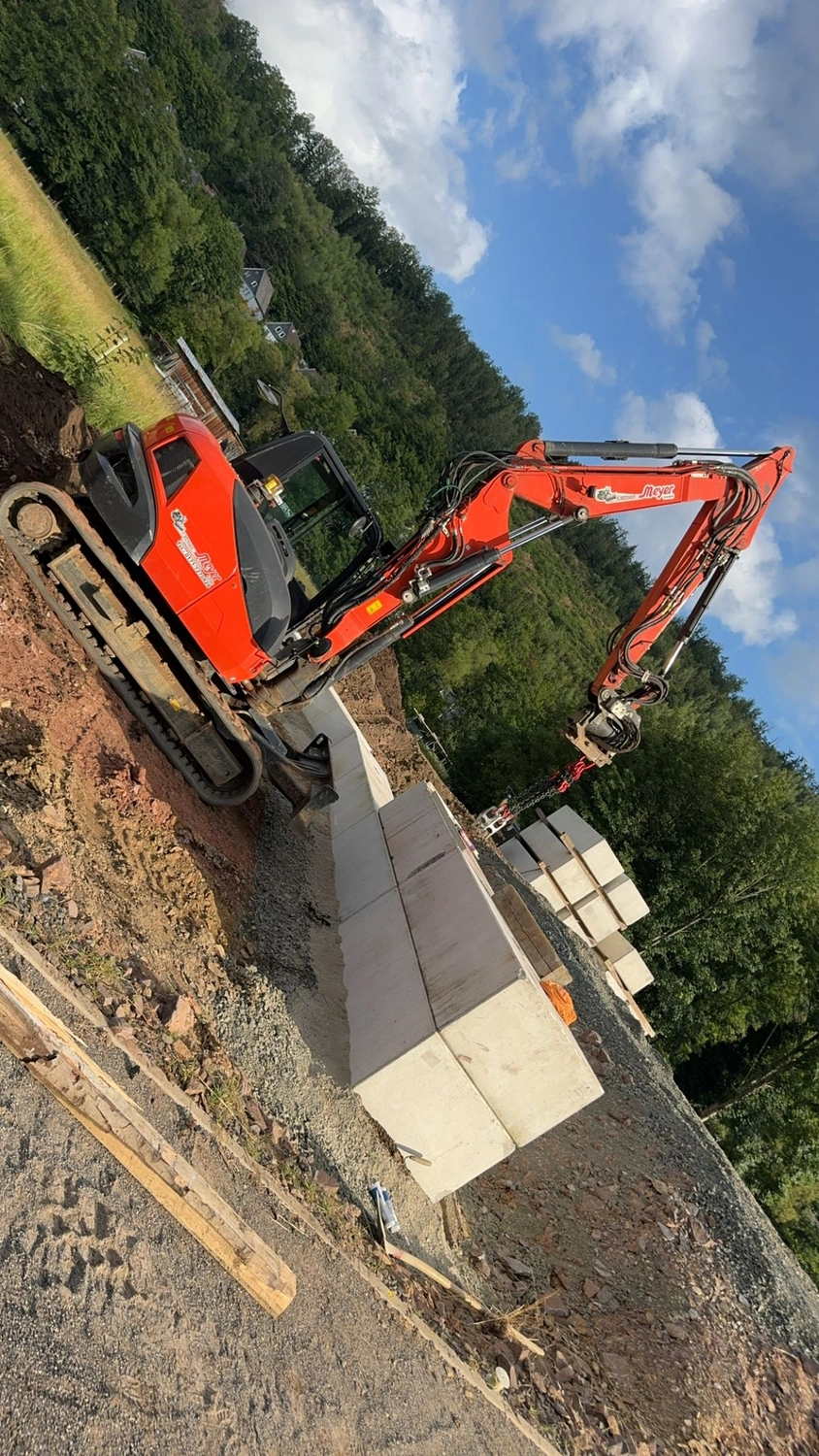 Hans Werner Meyer GmbH - Tiefbauunternehmen - Ein orangefarbener Kubota-Bagger stapelt große Betonblöcke, um auf einer abschüssigen Baustelle eine Stützmauer zu errichten.