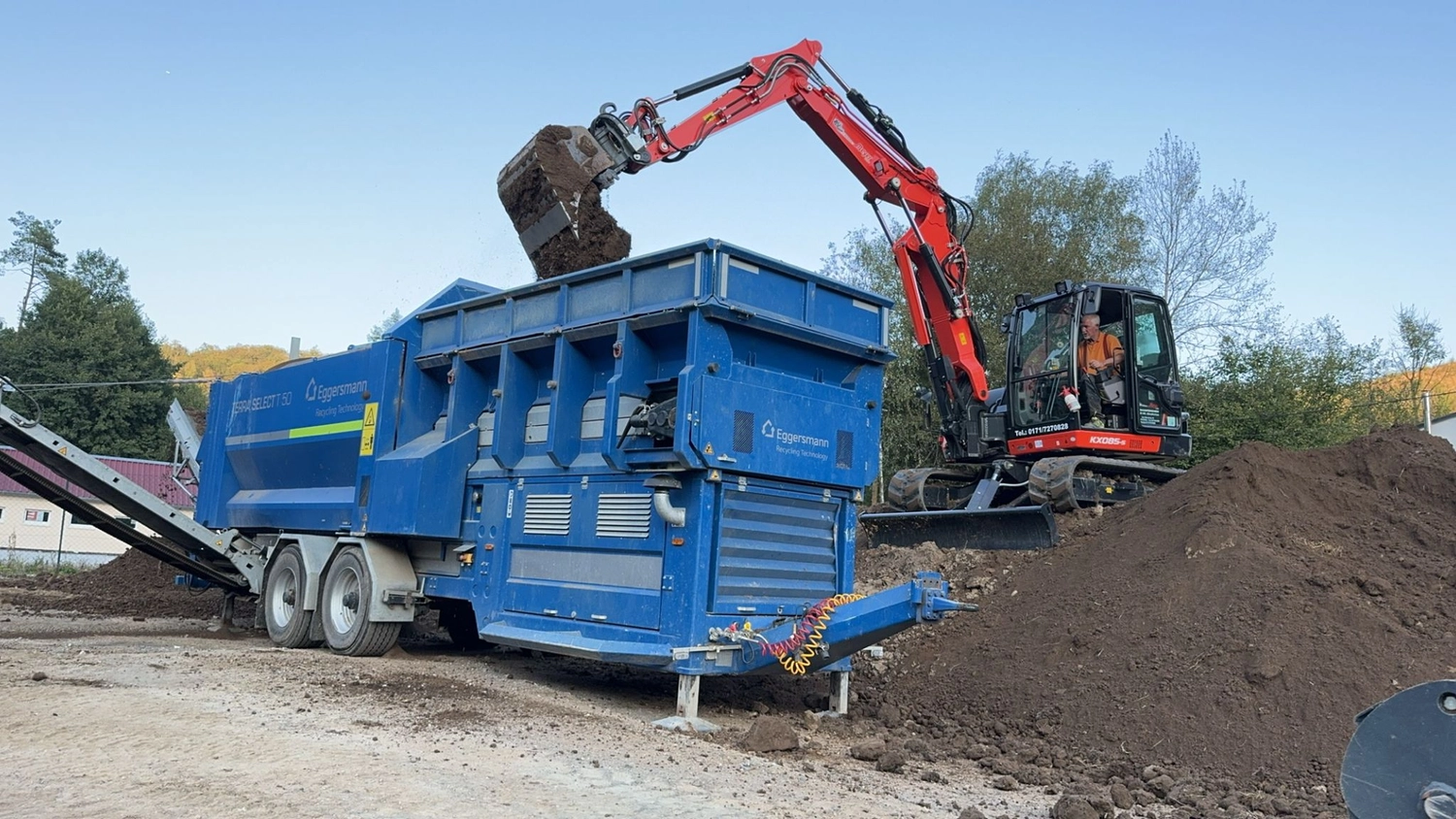 Hans Werner Meyer GmbH - Tiefbauunternehmen - Ein roter Bagger belädt eine große blaue Eggersmann-Trommelsiebmaschine auf einer Baustelle oder Recyclinganlage mit Erde.