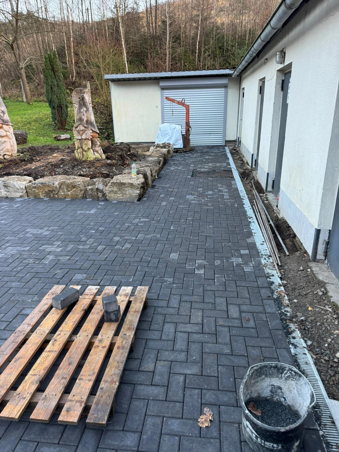 Hans Werner Meyer GmbH - Tiefbauunternehmen - Eine Weitwinkelaufnahme zeigt einen neu gepflasterten, aus dunkelgrauen, rechteckigen Ziegeln im Fischgrätmuster bestehenden Gehweg, der zu einem weißen Gebäude mit silbernem Rolltor führt. Im Vordergrund steht eine Holzpalette auf den Pflastersteinen, rechts ist ein Eimer mit Kies zu sehen.