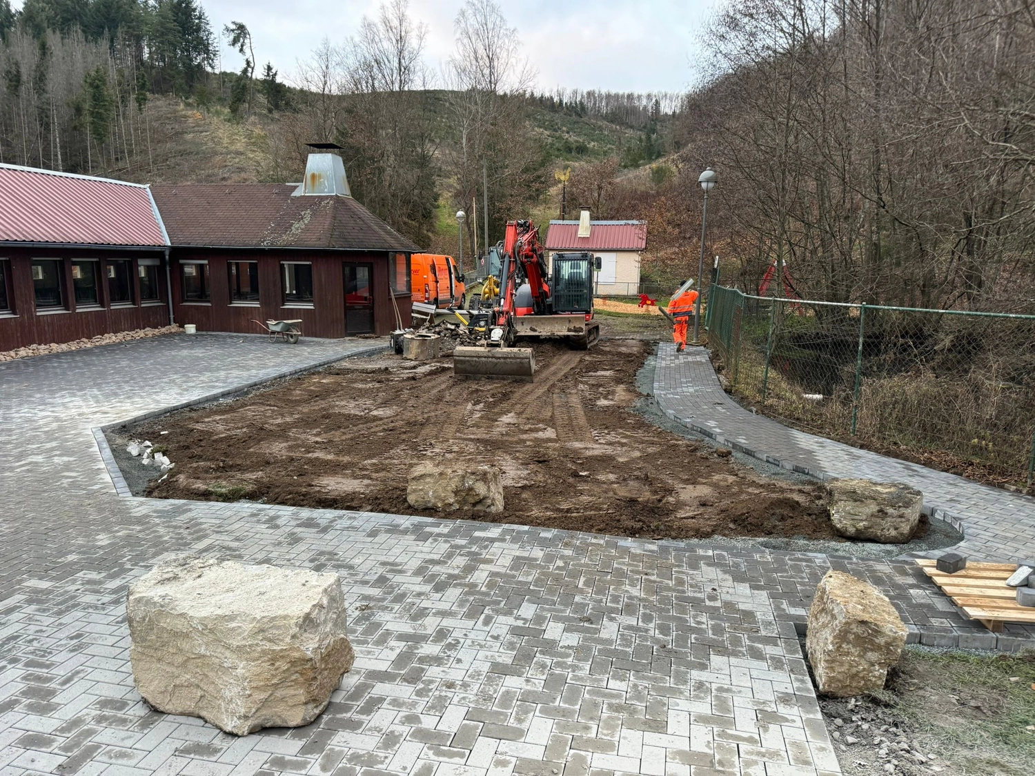 Hans Werner Meyer GmbH - Tiefbauunternehmen - Baustelle auf einem gepflasterten Platz mit einem kleinen roten Bagger, großen Steinen und einem Gebäude im Hintergrund.
