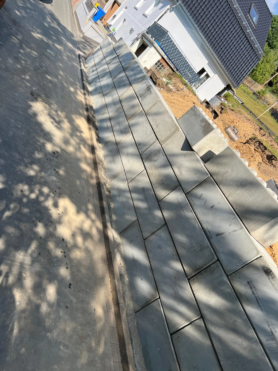Hans Werner Meyer GmbH - Tiefbauunternehmen - Eine Außenaufnahme aus der Vogelperspektive zeigt eine neu errichtete Stützmauer aus großen, rechteckigen grauen Betonblöcken, die entlang einer gepflasterten Straße in der Nähe eines weißen Hauses mit dunklem Ziegeldach terrassenförmig angelegt ist.