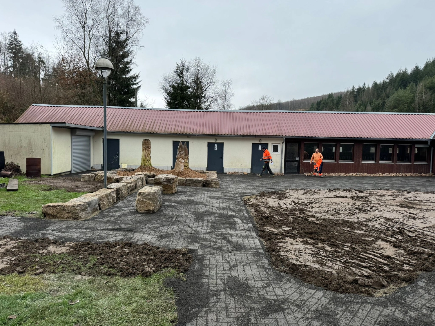 Hans Werner Meyer GmbH - Tiefbauunternehmen - Bauarbeiter in Warnwesten arbeiten in der Nähe eines niedrigen, weißen Gebäudes mit rotem Metalldach und mehreren Türen, das von großen Ziersteinen und einem teilweise mit Schlamm bedeckten gepflasterten Innenhof umgeben ist.