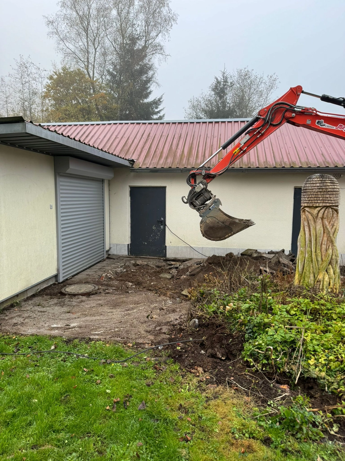 Hans Werner Meyer GmbH - Tiefbauunternehmen - Ein orangefarbener Baggerarm gräbt in ein Erdloch zwischen einem Gebäude und einem Garten. Im Vordergrund sind ein Rolltor, eine dunkle Eingangstür und eine dekorative Holzschnitzerei zu sehen.