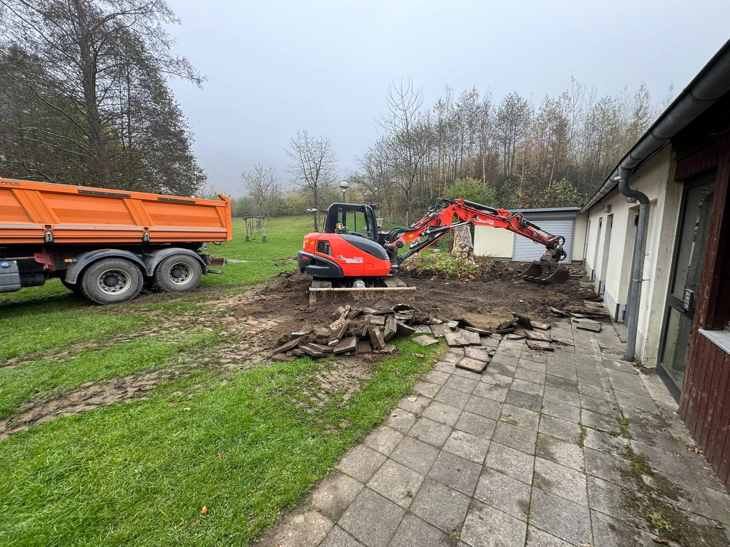 Hans Werner Meyer GmbH - Tiefbauunternehmen - Ein orangefarbener Kompaktbagger steht auf einem unbefestigten Stück Land neben einem Gebäude, nachdem er Steinpflaster ausgegraben hat, während ein orangefarbener Muldenkipper in der Nähe auf dem Rasen geparkt ist.