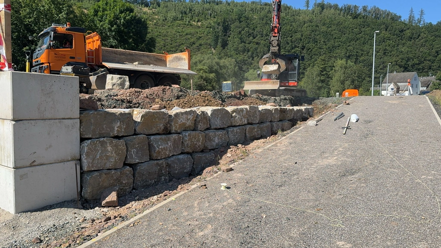 Hans Werner Meyer GmbH - Tiefbauunternehmen - Ein orangefarbener Muldenkipper und ein Bagger errichten eine Steinstützmauer entlang einer asphaltierten Straße in einem hügeligen, bewaldeten Gebiet.