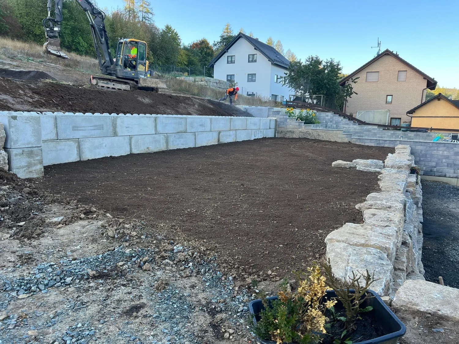 Hans Werner Meyer GmbH - Tiefbauunternehmen - Ein Bagger steht auf einem Hang hinter einer gestuften Stützmauer aus großen Betonblöcken und Naturstein und überblickt eine frisch eingeebnete Fläche in einem Wohngarten.