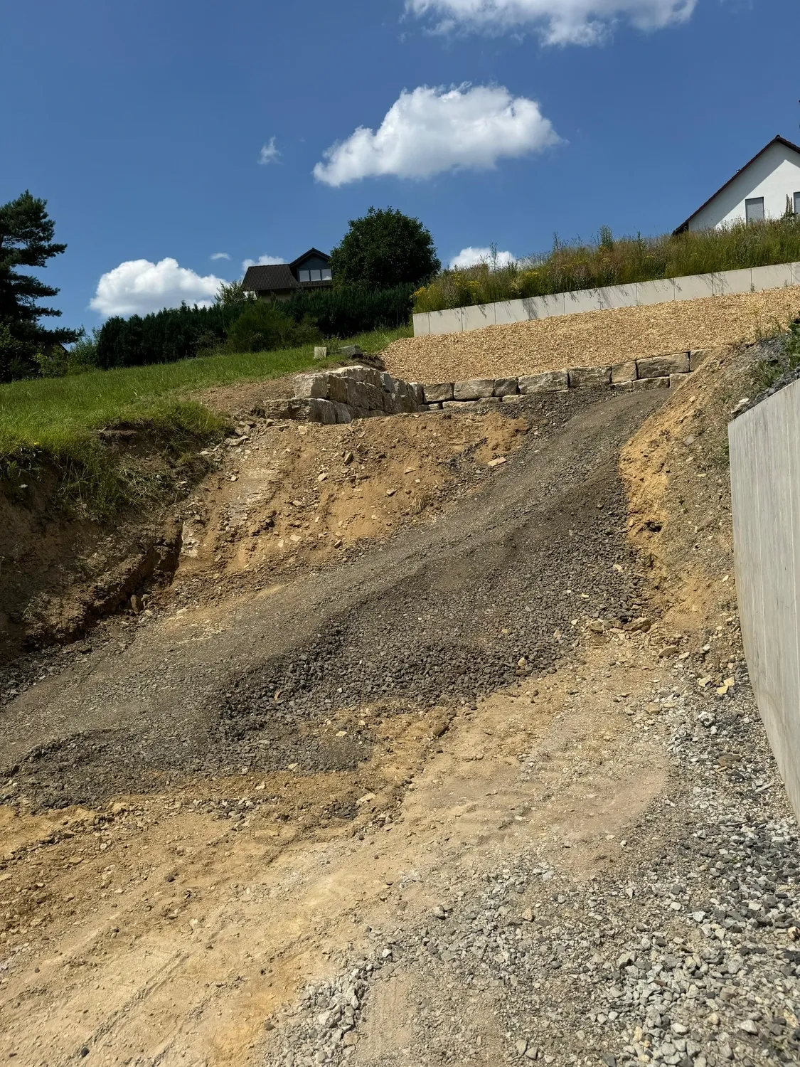 Hans Werner Meyer GmbH - Tiefbauunternehmen - Ein steiler, unbefestigter Schotterhang auf einem Wohngrundstück mit einer gestapelten Steinmauer am oberen Ende und einer Betonmauer rechts.