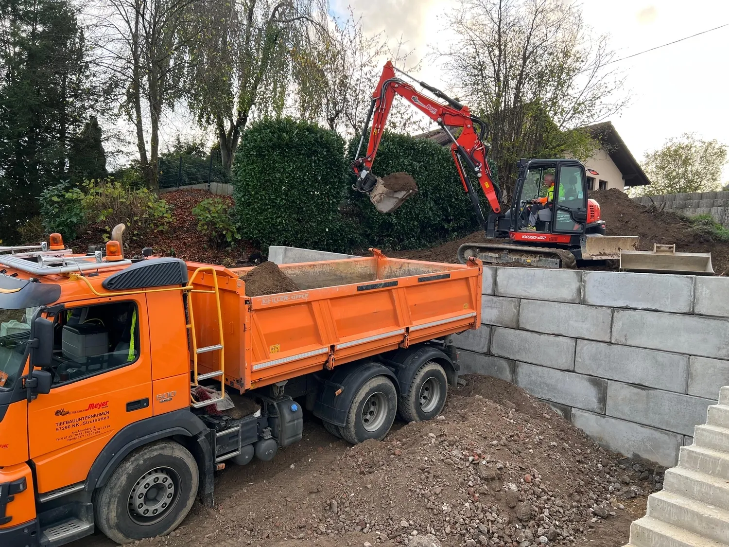 Hans Werner Meyer GmbH - Tiefbauunternehmen - Ein orangefarbener Muldenkipper wird auf einer Baustelle mit einer Stützmauer aus Betonblöcken von einem roten Minibagger mit Erde beladen.