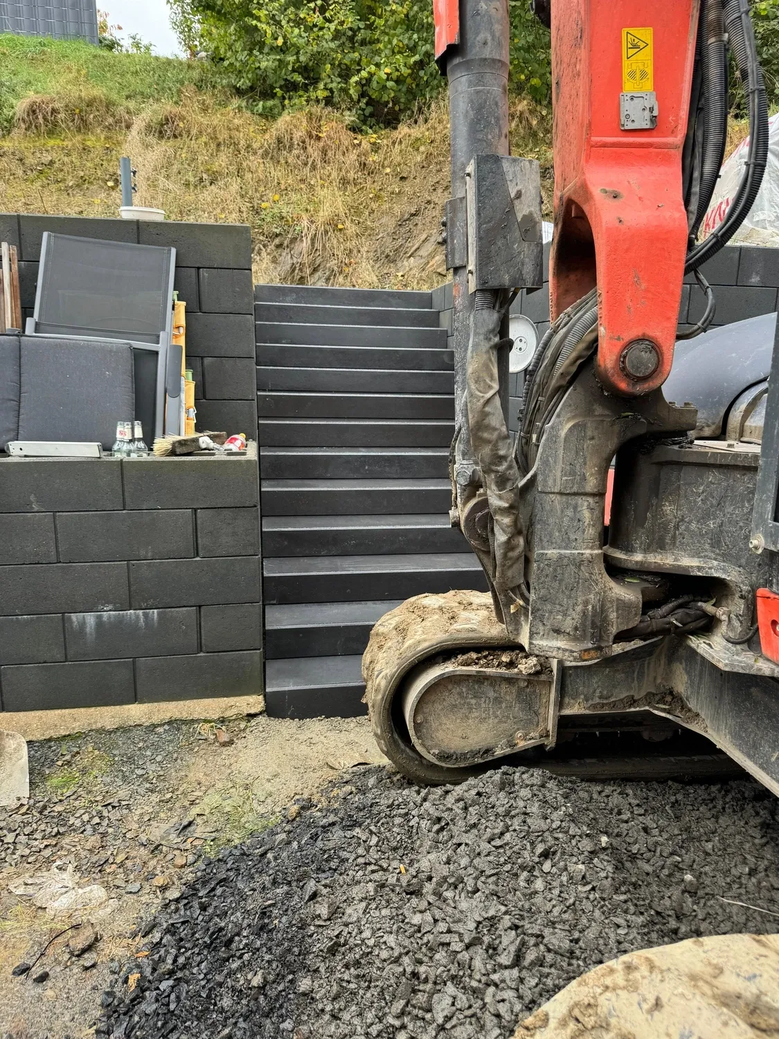 Hans Werner Meyer GmbH - Tiefbauunternehmen - Ein Bagger parkte auf einer Kiesfläche neben einer neu errichteten dunkelgrauen Steintreppe und einer dazu passenden Stützmauer.