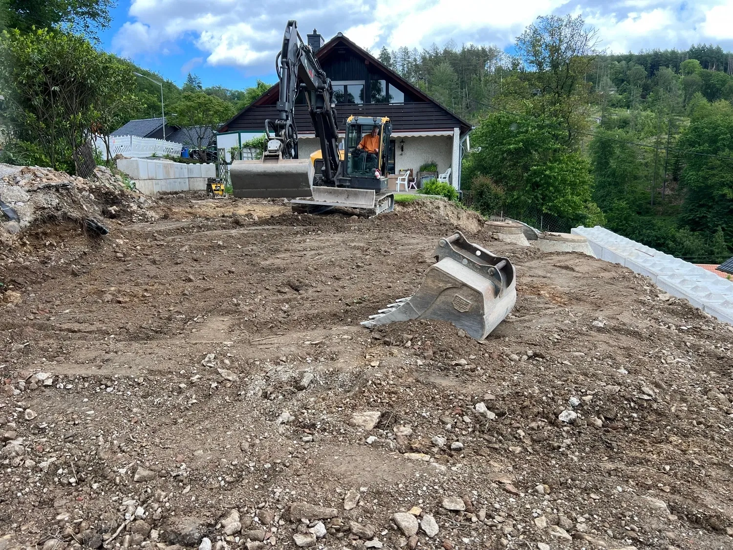 Hans Werner Meyer GmbH - Tiefbauunternehmen - Auf einer Baustelle vor einem Haus ist ein Bagger im Einsatz; im Vordergrund steht eine große Ersatzschaufel.