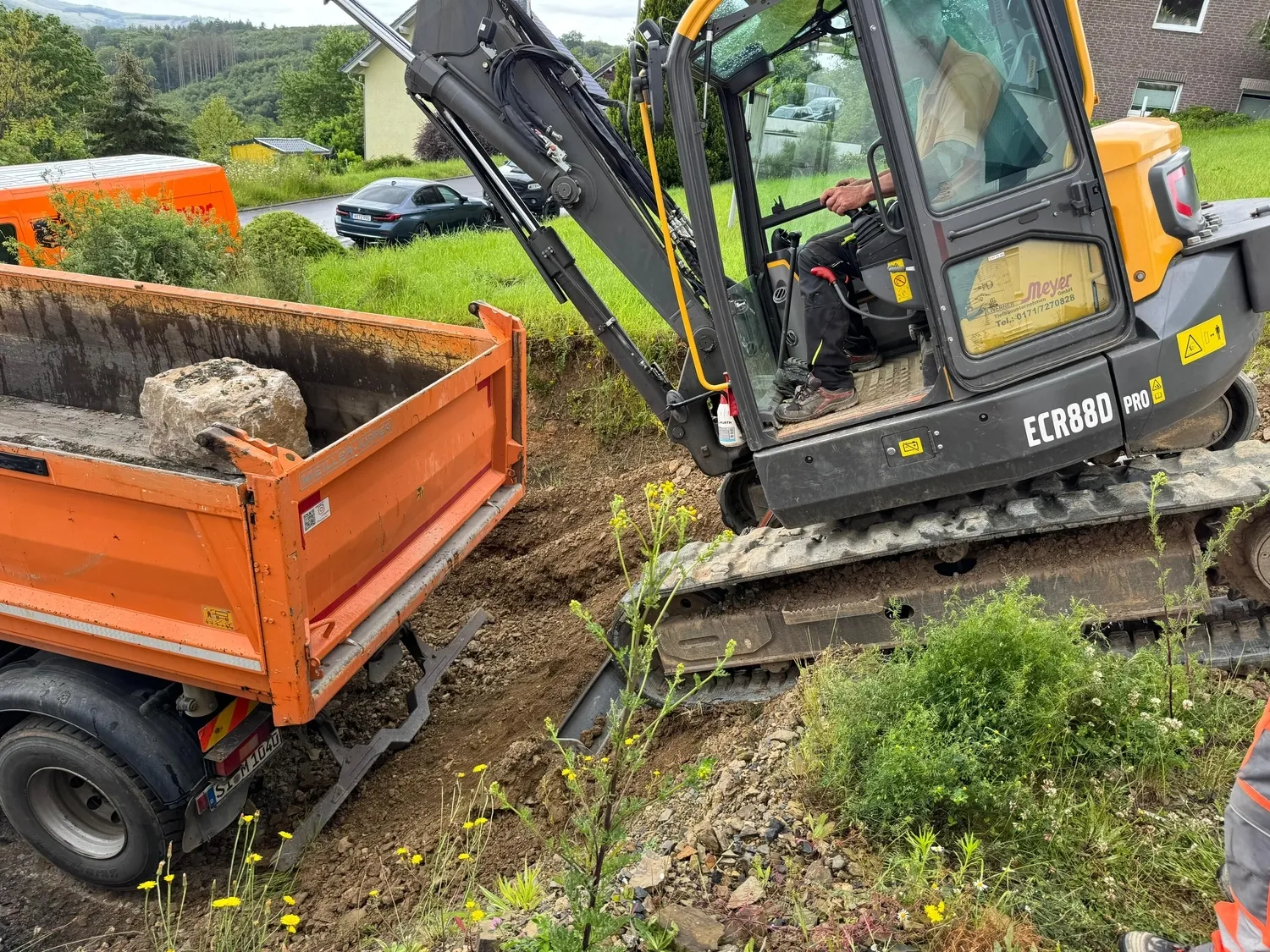 Hans Werner Meyer GmbH - Tiefbauunternehmen - Ein Volvo ECR88D Kompaktbagger verlädt schwere Steine ​​auf die Ladefläche eines orangefarbenen Muldenkippers auf einer Wohnbaustelle.