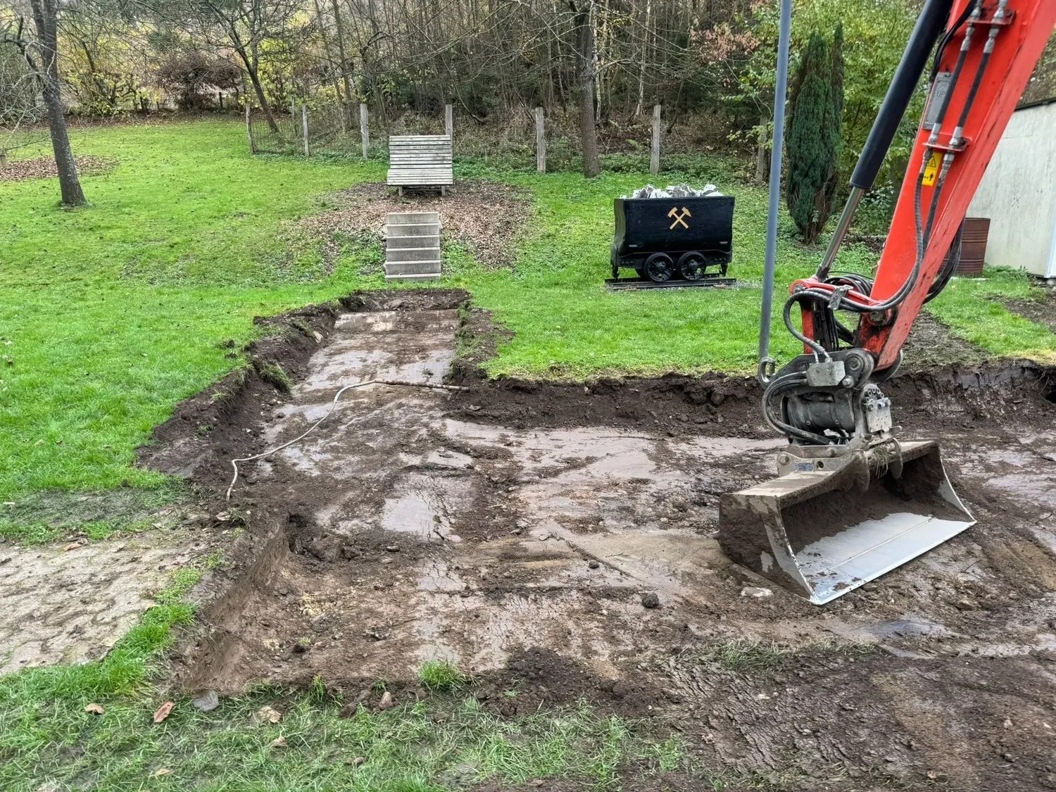 Hans Werner Meyer GmbH - Tiefbauunternehmen - Eine orangefarbene Baggerschaufel steht auf einer matschigen Stelle, die in einem grasbewachsenen Hinterhof gerade geräumt wird, neben einem schwarzen Lorenwagen.