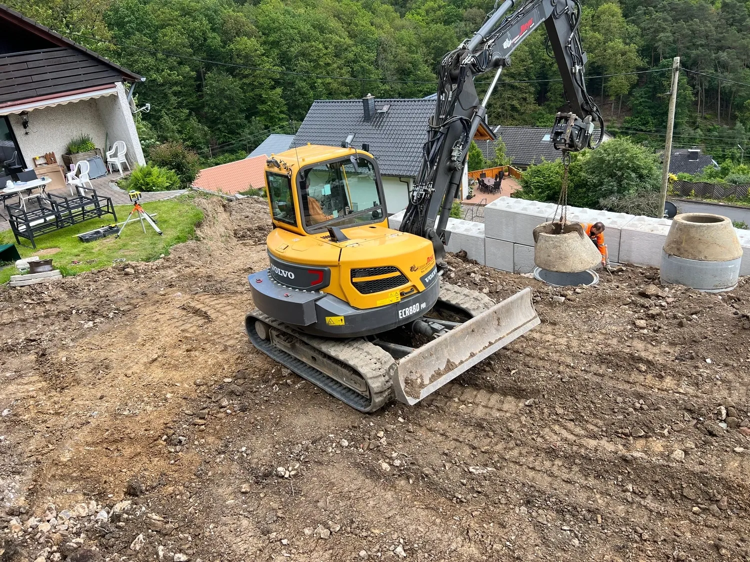 Hans Werner Meyer GmbH - Tiefbauunternehmen - Ein Volvo ECR88D Kompaktbagger ist auf einer Wohnbaustelle im Einsatz und hebt mit seinem Ausleger einen Betonring an, während im Hintergrund ein Arbeiter assistiert.