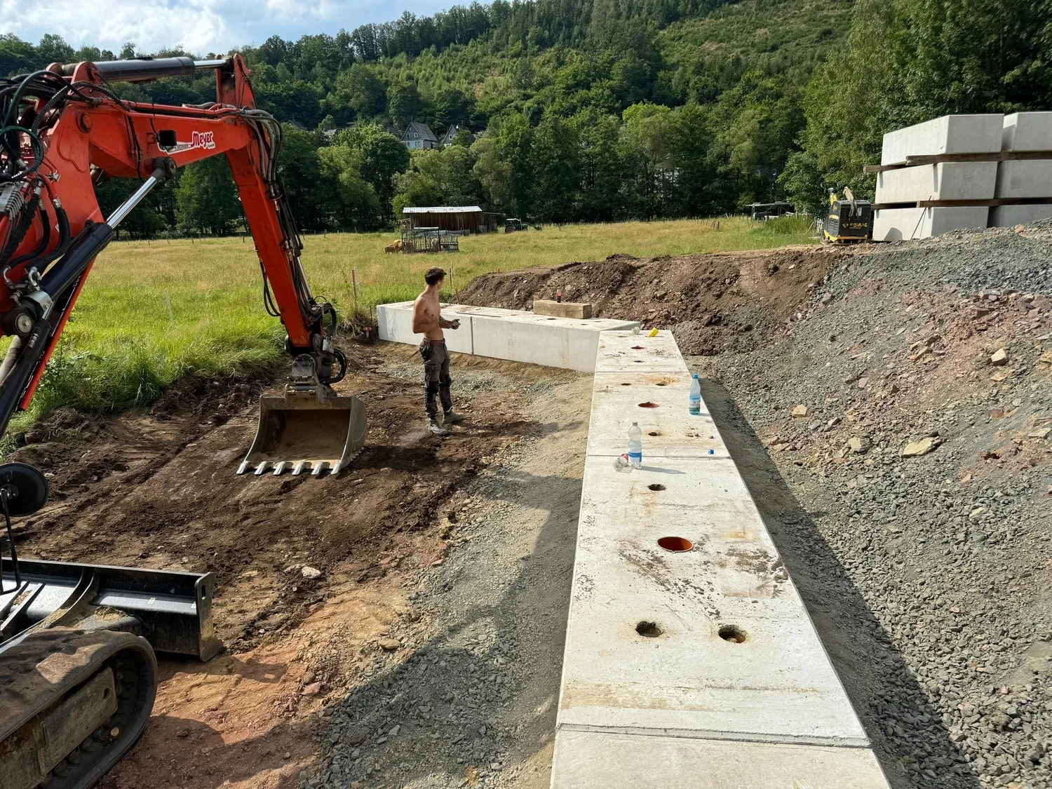 Hans Werner Meyer GmbH - Tiefbauunternehmen - Hans Werner Meyer GmbH - Tiefbauunternehmen - Ein orangefarbener Bagger arbeitet auf einer Baustelle neben einer neu errichteten L-förmigen Betonstützmauer in einer ländlichen, hügeligen Landschaft.